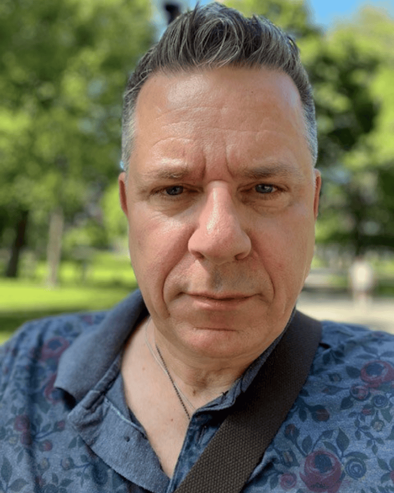 Close-up portrait of a man with short gray hair wearing a patterned polo shirt and shoulder strap, standing outdoors with trees in the background.